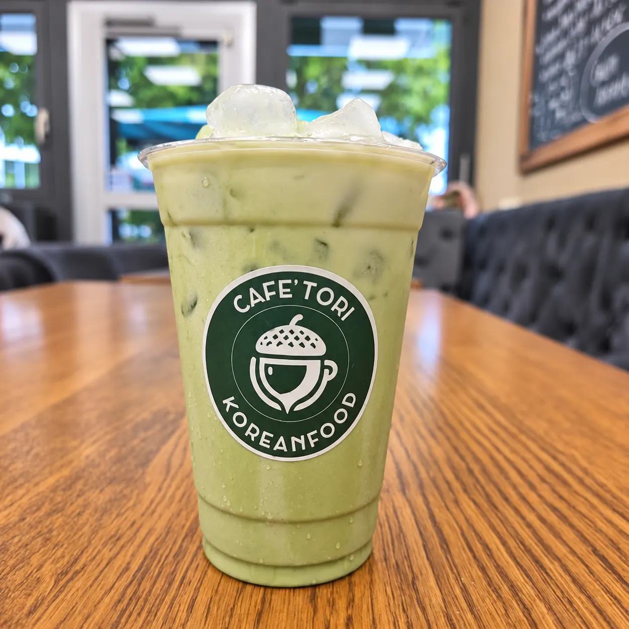 Iced Matcha Latte im Café-Tori-Becher mit Eichel-Logo auf einem Holztisch